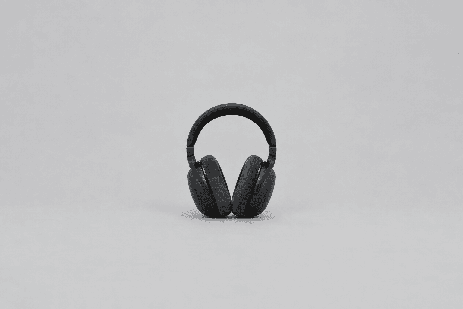 A plain black pair of over-ear studio headphones with dark grey fabric ear pads, standing upright and centred against a plain pale grey background with a soft shadow beneath
