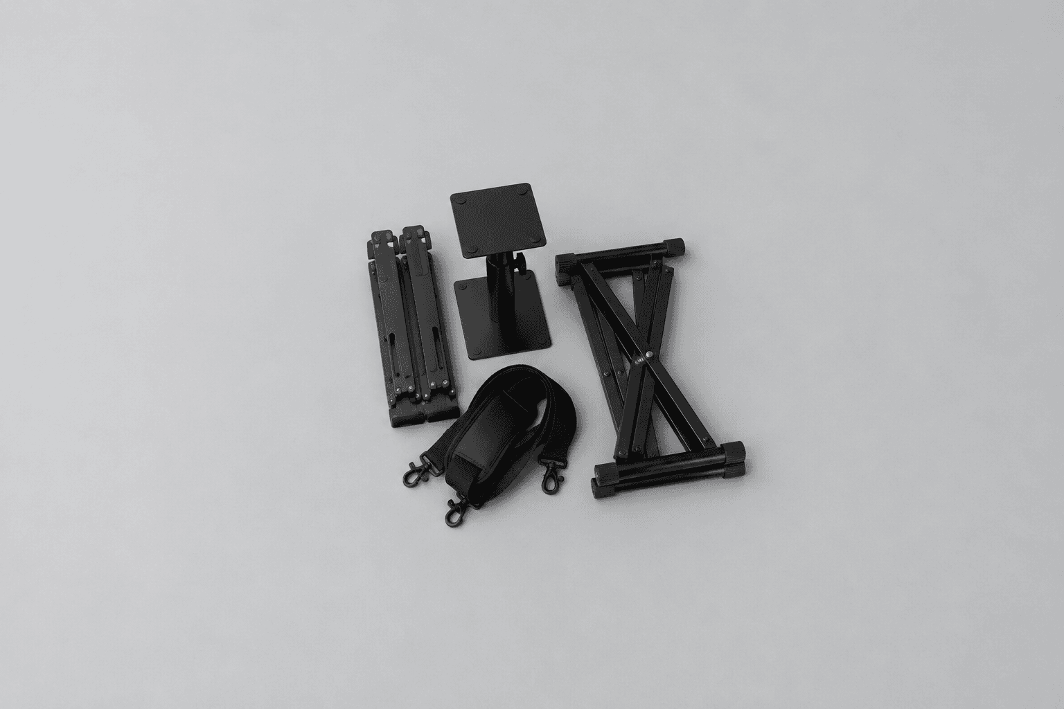 Overhead photograph of four black home studio accessories grouped in the centre of a plain pale grey surface: a folded metal laptop stand, a short desktop speaker stand with a square top plate, a padded shoulder strap with metal clips, and a folded double-braced X keyboard stand