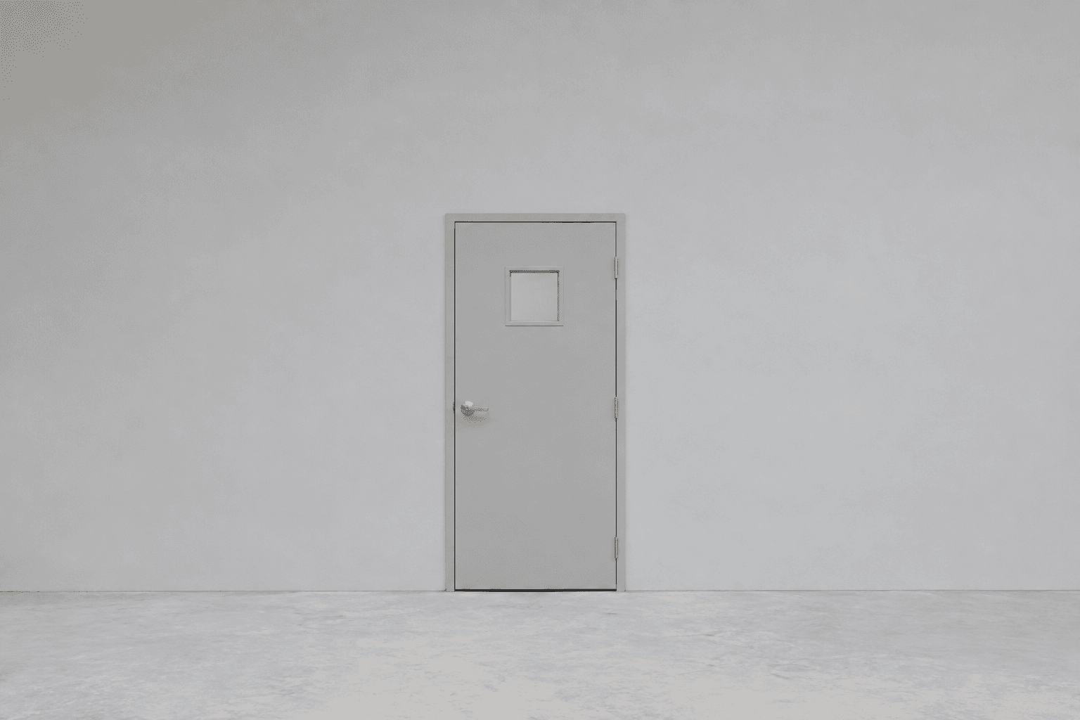 A single plain pale grey closed door with a small square window and a brushed metal lever handle, centred in a wide empty pale grey wall above a bare concrete floor, with large areas of empty space on both sides