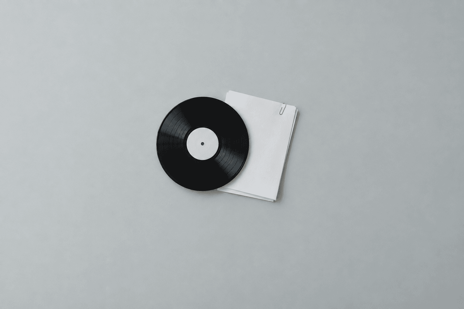 A black vinyl record with a blank cream centre label lying across a small stack of blank white paper held with a silver paperclip, shot from directly above in the centre of a wide empty pale grey desk