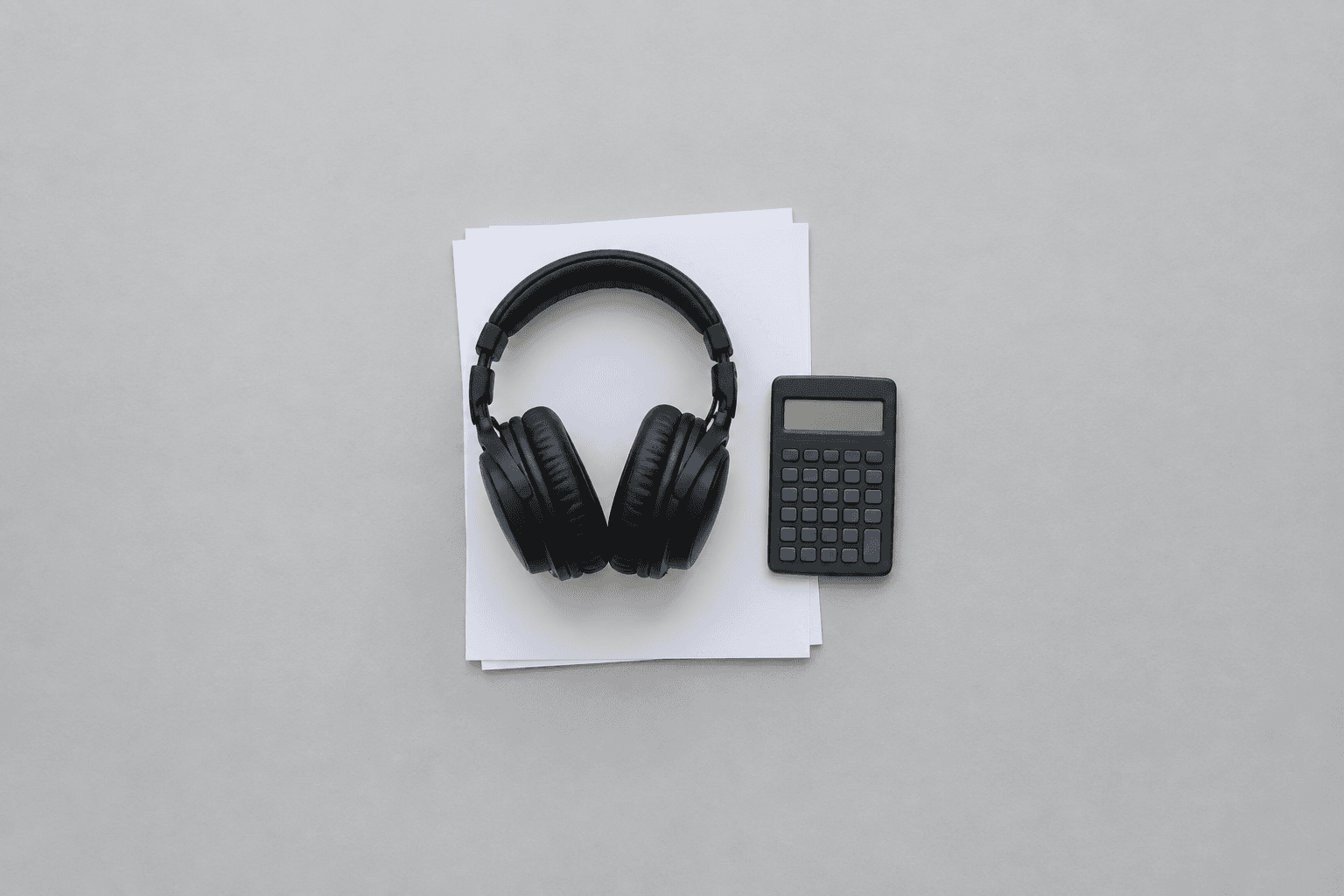 A pair of black closed-back studio headphones resting on a small stack of blank white paper next to a plain pocket calculator, photographed from directly above as one tight cluster in the centre of a wide empty pale grey desk
