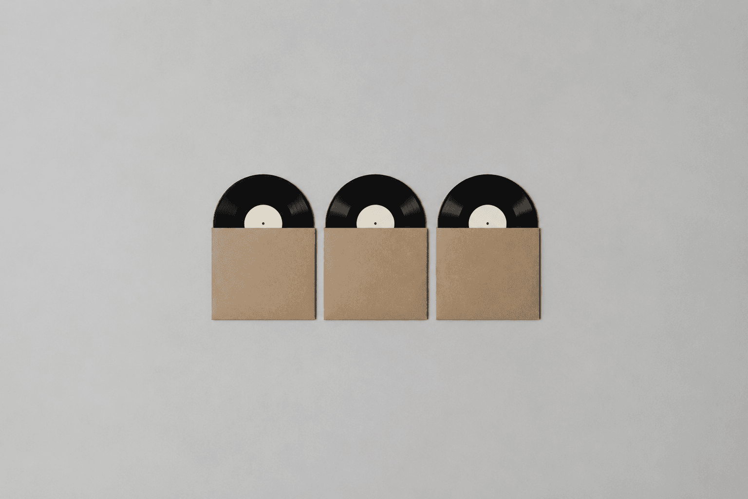 Three plain black vinyl records partly slid out of unprinted brown cardboard sleeves, laid in a row and photographed from above in the centre of a wide empty pale grey surface