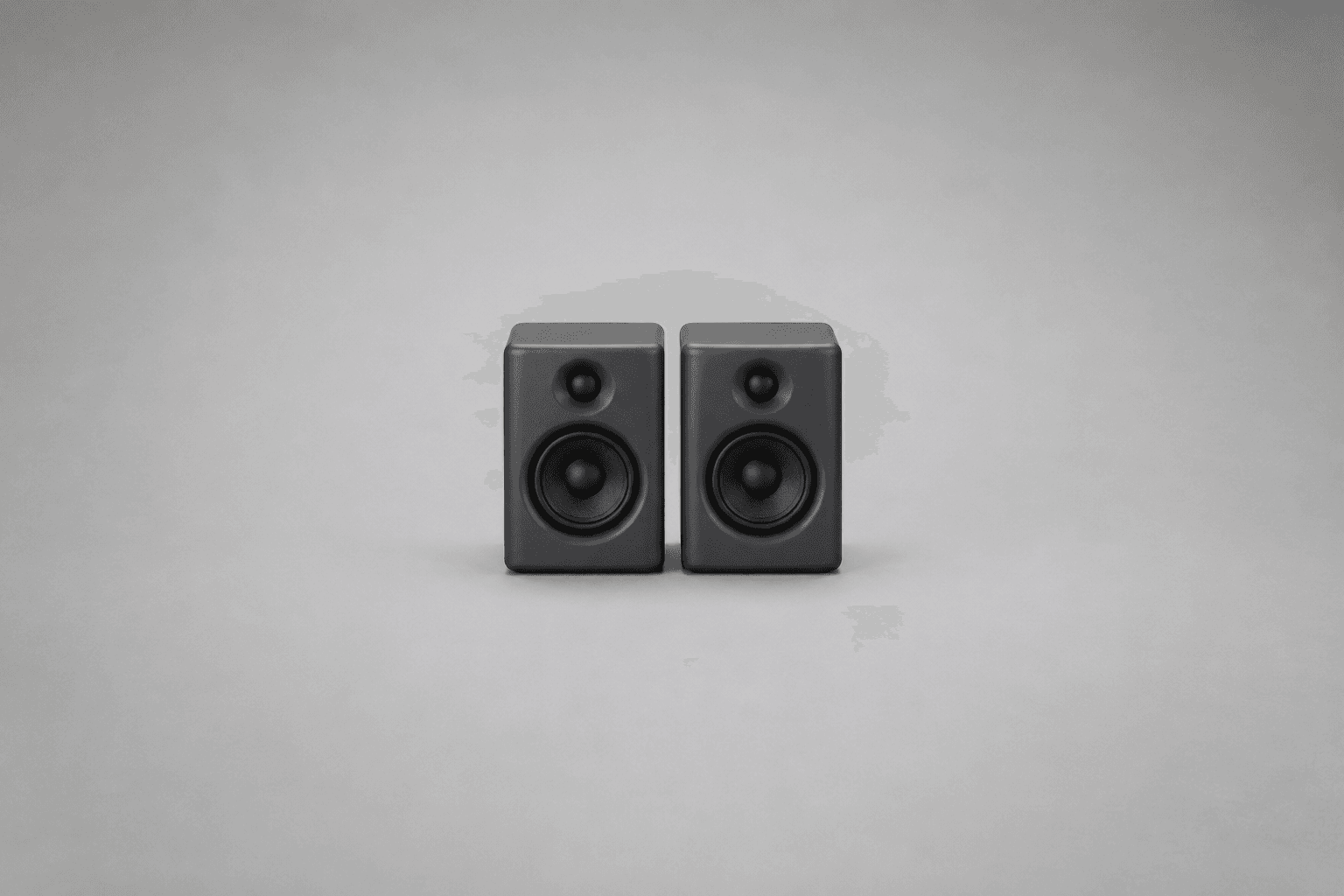 Two identical matte dark-grey studio monitor speakers standing side by side with a narrow gap between them, small and centred on a plain unbroken mid-grey background with wide empty space all around