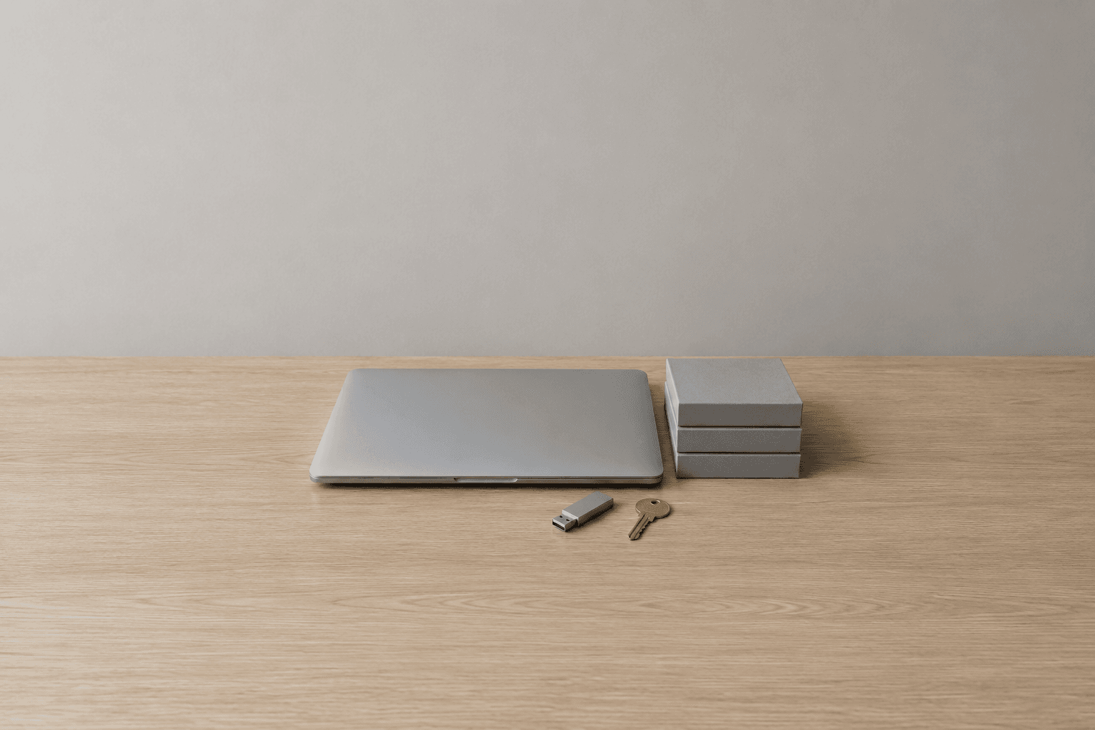 A closed silver laptop, a stack of three plain grey boxes, a small USB stick and a brass key grouped in the centre of a pale oak desk against a plain grey wall