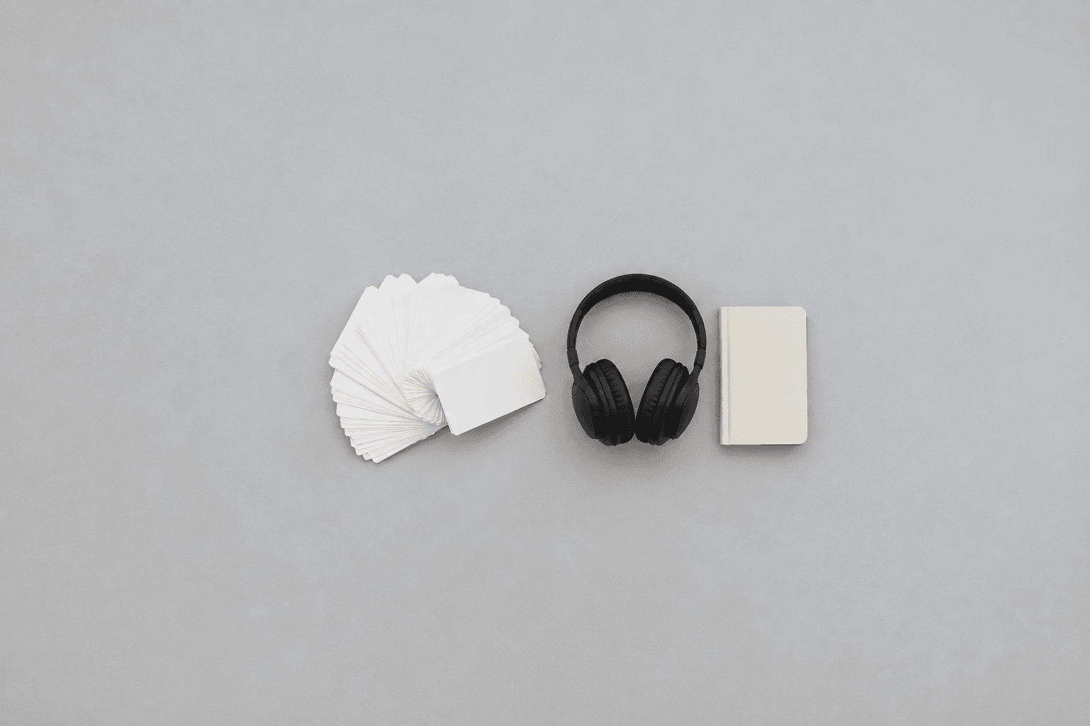 A fanned-out pile of blank white index cards, a pair of black over-ear headphones and a small blank cream notebook laid in a row, shot from directly above in the centre of a wide empty pale grey desk