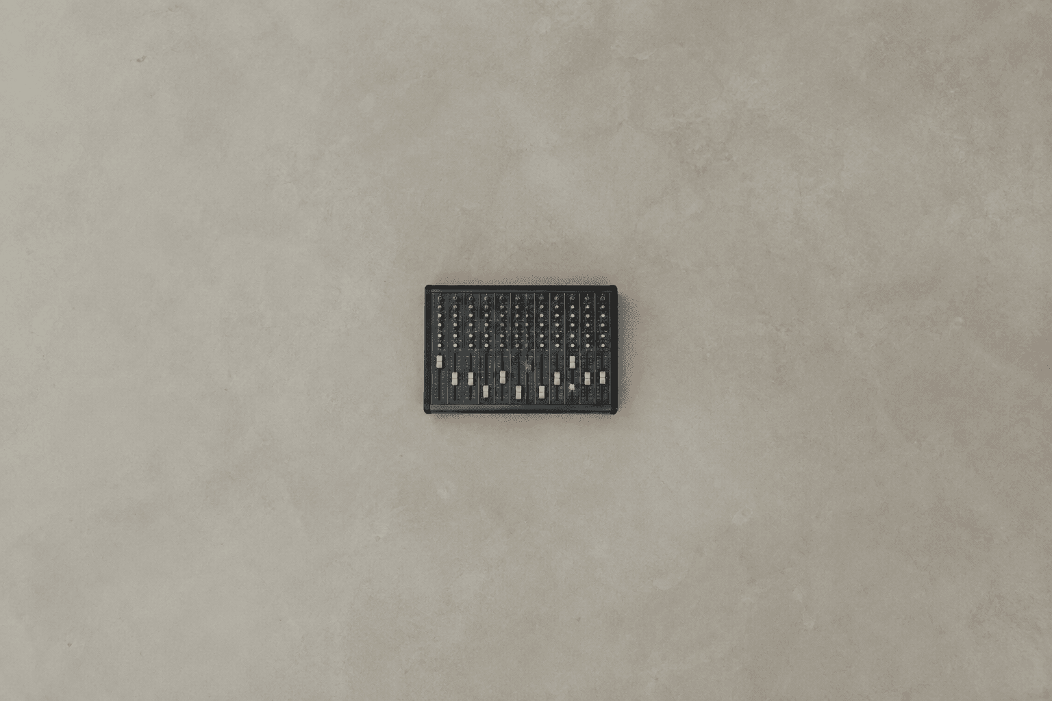 A small black analogue mixing console, its row of channel faders left at staggered heights, photographed from directly above and sitting alone in the centre of a large empty expanse of pale grey concrete floor