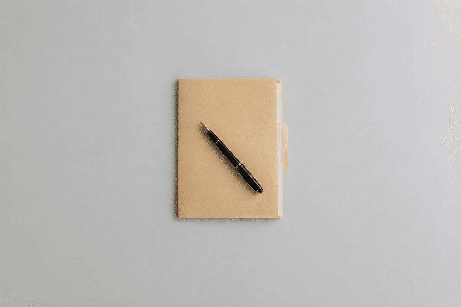 A closed kraft-brown document folder with a black and gold fountain pen resting diagonally on top of it, photographed from directly above in the centre of a wide empty pale grey desk with generous margin on all sides