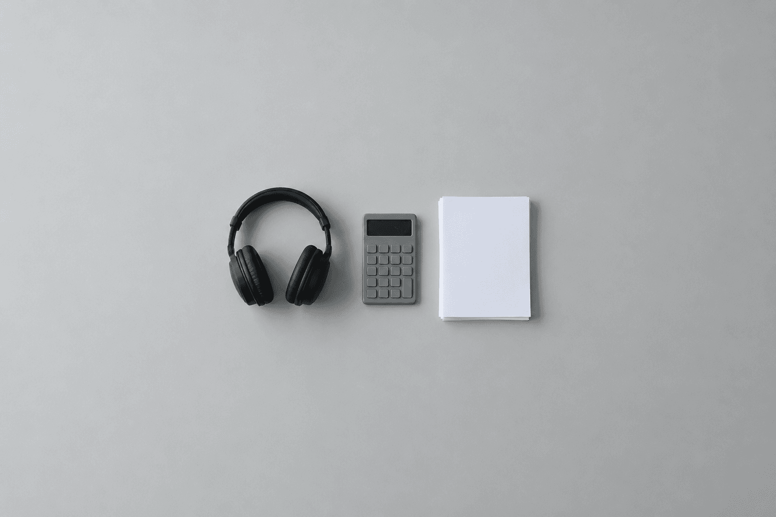 A pair of black closed-back studio headphones, a plain grey pocket calculator with a blank display and a neat stack of blank white paper, laid in a row at the centre of a wide plain grey surface and shot from above